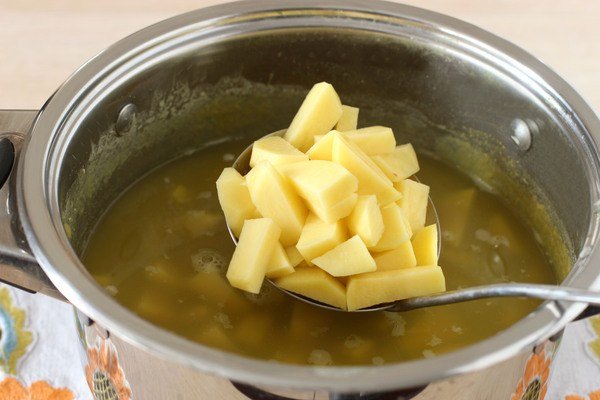 Нарезанный маленькими кусочками сырой картофель в большой ложке над кастрюлей с супом