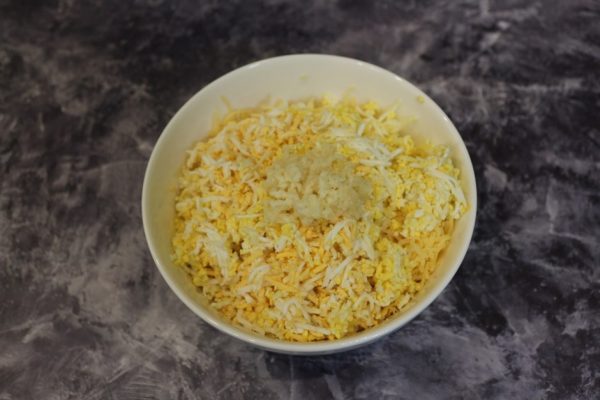 Тёртые варёные яйца, твёрдый сыр и чеснок в белой миске на столе
