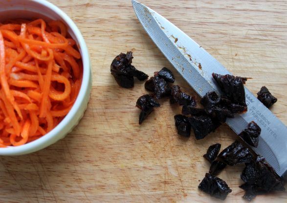 нарезанный кусочками чернослив, нож и морковь по-корейски в миске на столе