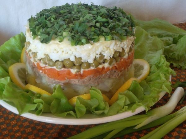 Слоёный салат из печени трески и консервированного зелёного горошка на тарелке с листьями салата и дольками лимона
