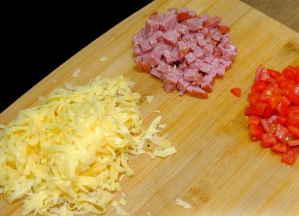 Тёртый твёрдый сыр и нарезанные мелкими кубиками свежие помидоры с варёной колбасой на деревянной разделочной доске